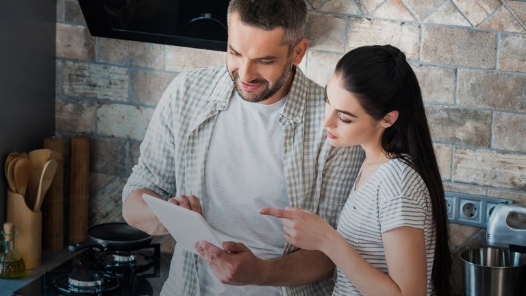 A man and a woman stand in the kitchen looking at a tablet